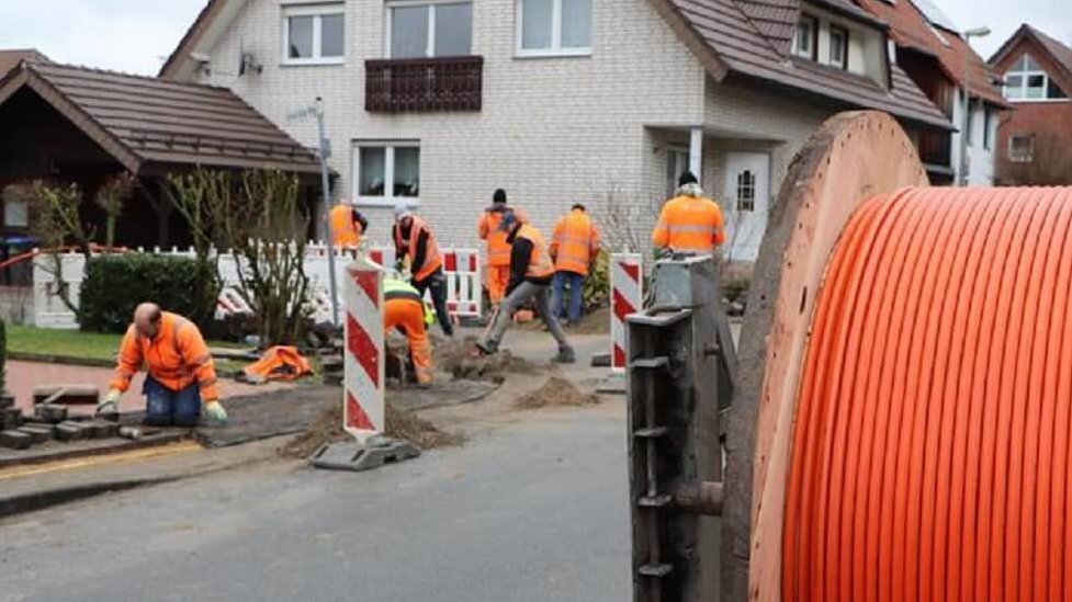 Viele Bauarbeiter vor einen weißen Klinkerhaus, im Vordergrund ein große Rolle mit orangefarbener Leitung